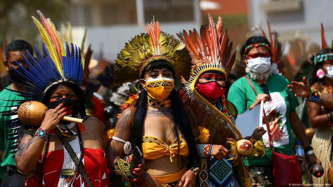 Mulheres indígenas com cocares e vestimentas tradicionais. Ela usam máscara de proteção