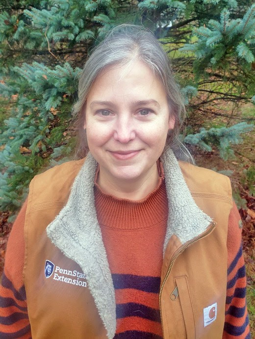 A woman stands outdoors in front of an evergreen tree. She wears a vest with the text: Penn State Extension