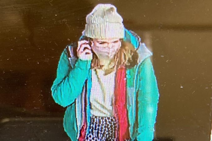 A screenshot of a woman in a beanie, face mask and green coat walking while speaking on the phone