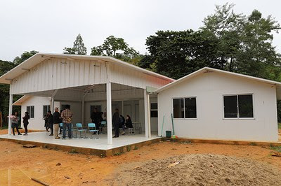 A escola da aldeia indígena. Um prédio em alvenaria e madeira, pintado de branco. No centro, se vê uma área externa coberta, onde pessoas conversam. Em ambos os lados dessa área, se veem as fachadas das salas de aula, que são duas construções menores com duas janelas cada uma.