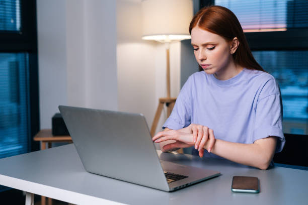 Overworked redhead young woman typing on laptop actively feeling pain in wrist sitting at desk near window in evening. Overworked redhead young woman typing on laptop actively feeling pain in wrist sitting at desk near window in evening. Lady working on laptop and suffering from pain in hand, carpal tunnel syndrome. dark joint and muscle pain stock pictures, royalty-free photos & images