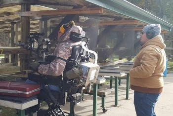 A person bundled in warm hunting camo clothing and sitting in a mobility chair operates an air-puff gun at a DNR shooting range.