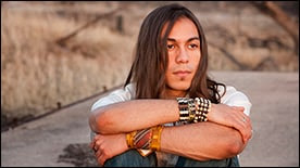 The figure shows a young adult American Indian male sitting outside and looking off into the distance.