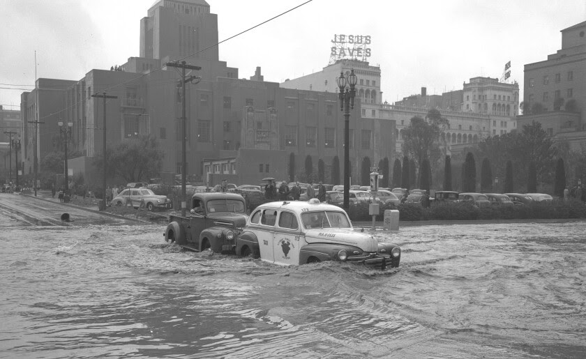 A truck pushes a car through deep water in a city intersection. Behind, a sign atop a building says "Jesus Saves."