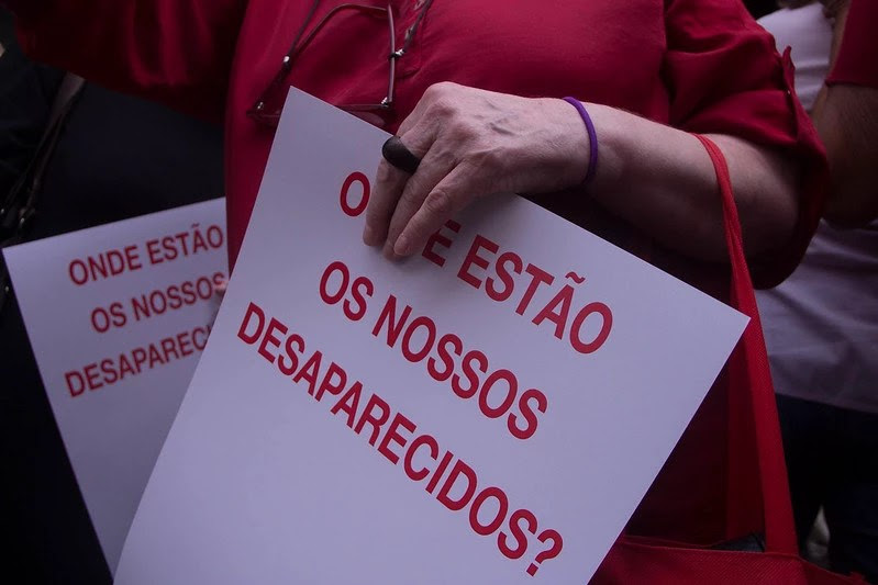 Foto com foco nas mãos de uma mulher durante ato Ditadura Nunca Mais, segurando um cartaz branco com a seguinte frase em vermelho: "onde estão os nossos desaparecidos?"