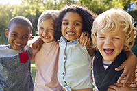 Photograph of a group of four diverse children approximately age 4 smiling at the camera with trees in the background.