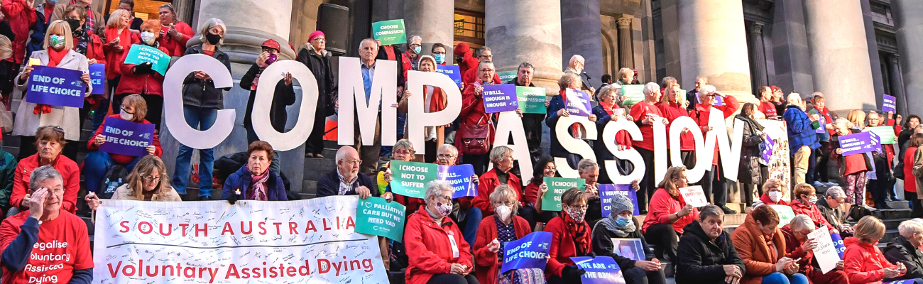 A crowd of people sit outside South Australia parliament house to support voluntary assisted dying. They hold a large sign that reads out 'compassion'
