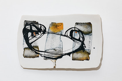 A photograph of a ceramics plate by Jennifer Alford. The object is white with black, grey, brown and yellow abstract drawing on, and it is displayed against a solid white background. 