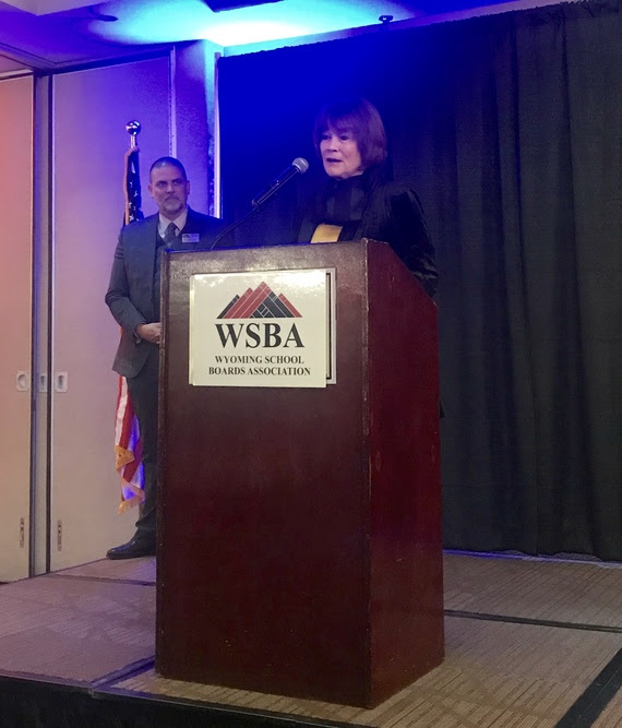 Judy speaks from a podium with a WSBA (Wyoming School Boards Association) sign on the front while WSBA Executive Director Brian Farmer observes.