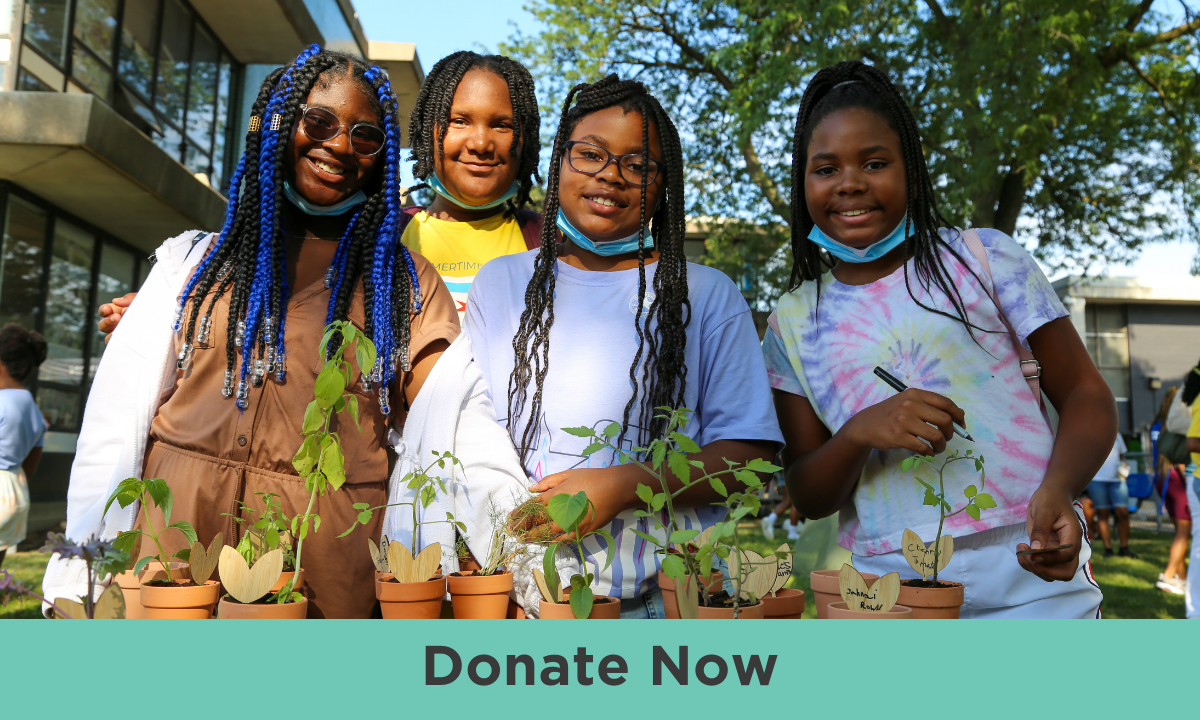 Four young girls of a range of medium deep skin tones smile to camera. They have their hair in braids of different lengths and colors, and wear an array of colorful shirts. All four have blue masks resting under their chins. On a table in front of them lie an assortment of small plants in terracotta pots.
