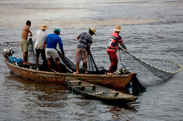 A imagem mostra alguns pescadores em cima de um pequeno barco estendendo a rede no mar