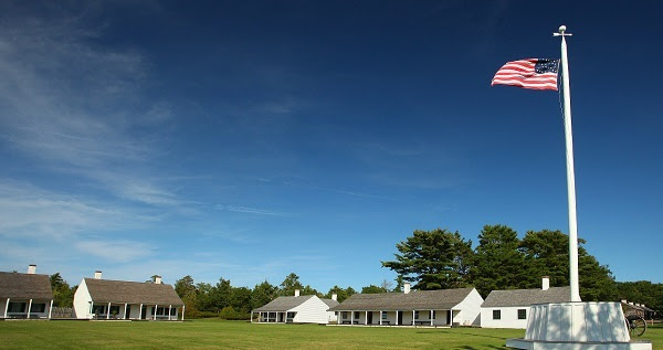 The U.S. flag billows to the left at the top of a tall white flagpole, over several low-lying white buildings with brown roofs; blue sky above