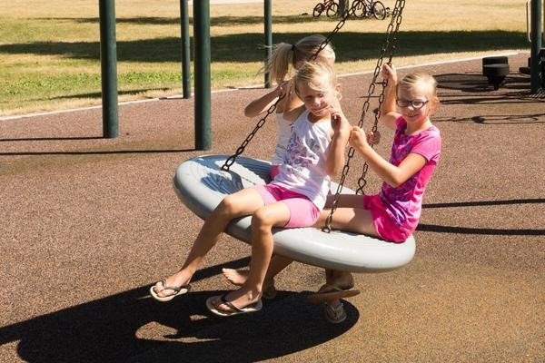 three little blond girls, dressed in pink and white T-shirts and shorts, hold onto the chains of a round swing as they spin around on a playground