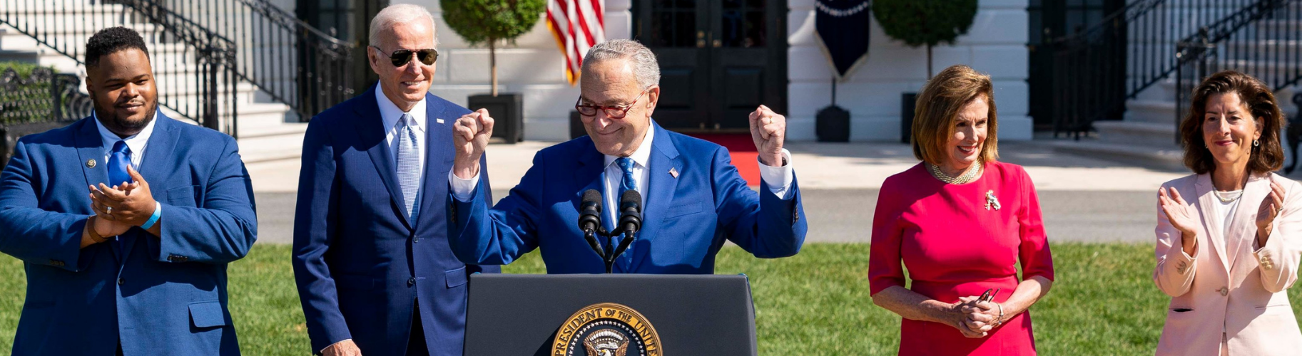 Senator Chuck Schumer stands at the podium with his arms raised in celebration and a smile. Behind him is businessman Joshua Aviv, President Joe Biden, Speaker Nancy Pelosi, and Secretary of Commerce Gina Raimondo, who are smiling and clapping.