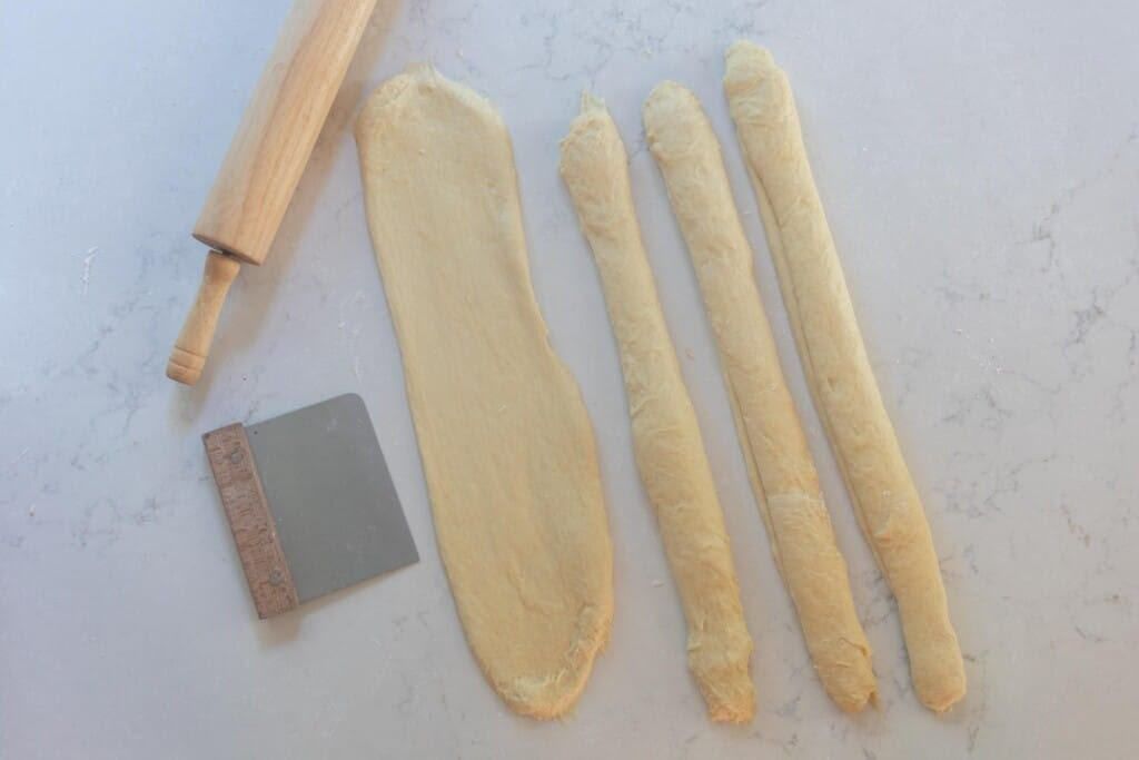 shaping long rolls of dough to braid on a white countertop with a rolling pin and bench scraper to the left