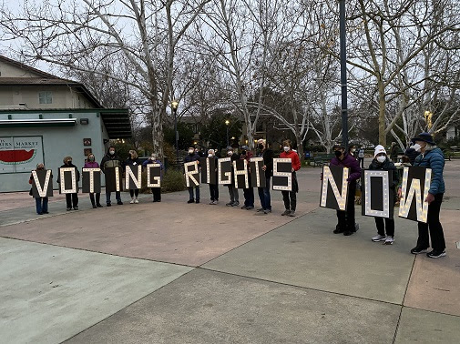 15 people stand in a line in Davis Central Park, each holding one large sign with a letter to spell out "Voting rights now"