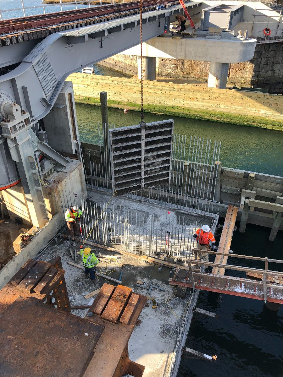 Two workers on an outside platform over the waterway and under the bridge place 6-foot sections of vertical rebar around a rectangular section of the platform. Another worker pulls a rope attached to a metal wall about 8 x 10 feet in size that is suspended over the platform on a chain. 