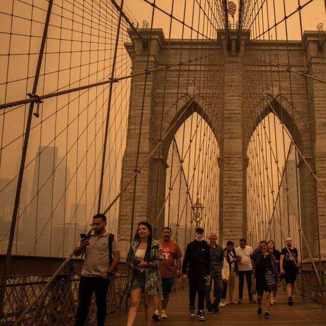 A dozen or so people walk amid the Brooklyn Bridge’s crisscrossing support wires and iconic stone arches, beneath a yellow-brown sky. A person in the foreground wears a shirt that reads “I (heart) NY.” 