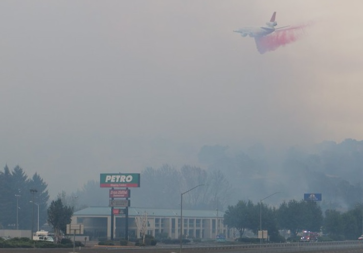 Image of a smoky town in Oregon due to wildfires. A fire-suppressant material drops from an airplane to fight the fire.