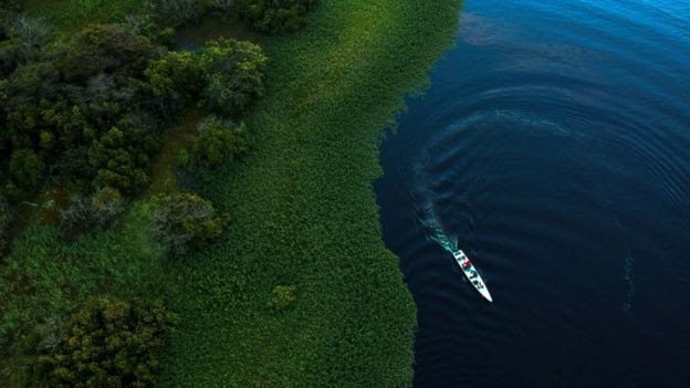 Foto mostra área de floresta e rio, pelo qual passa um pequeno barco, em vista aérea