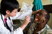 Woman doctor in white coat wears stethoscope, holds male patient's head back, uses pen light to look in his mouth