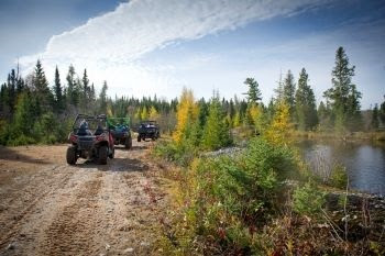 A single-file line of three off-road vehicles travel a dirt road in an area featuring conifer trees, a pond and blue, cloud-streaked sky