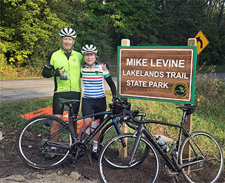 Two bicyclists pose for a photo in front of a sign for the Mike Levine Lakelands Trail State Park in southeast Michigan.