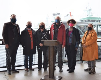 Photo of people standing for a photo behind a podium with a ferry in the background