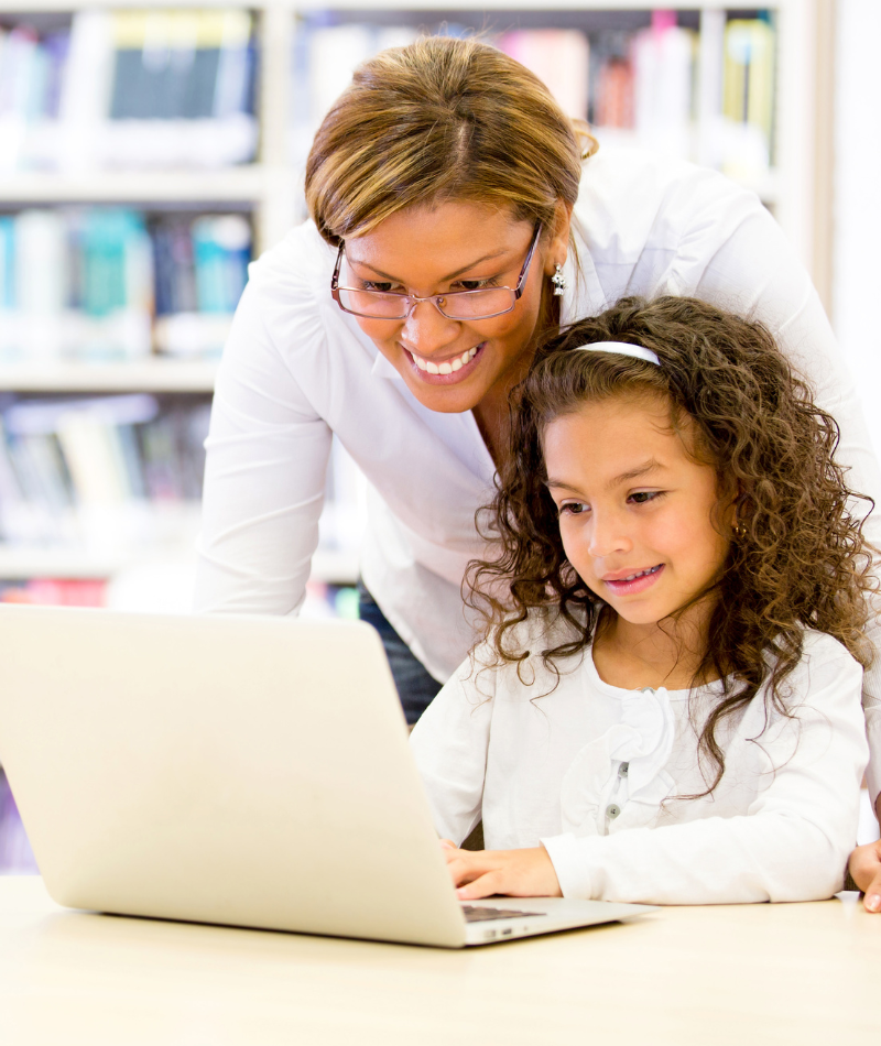 A young child sits in front of a laptop typing while a teacher looks over her shoulder smiling