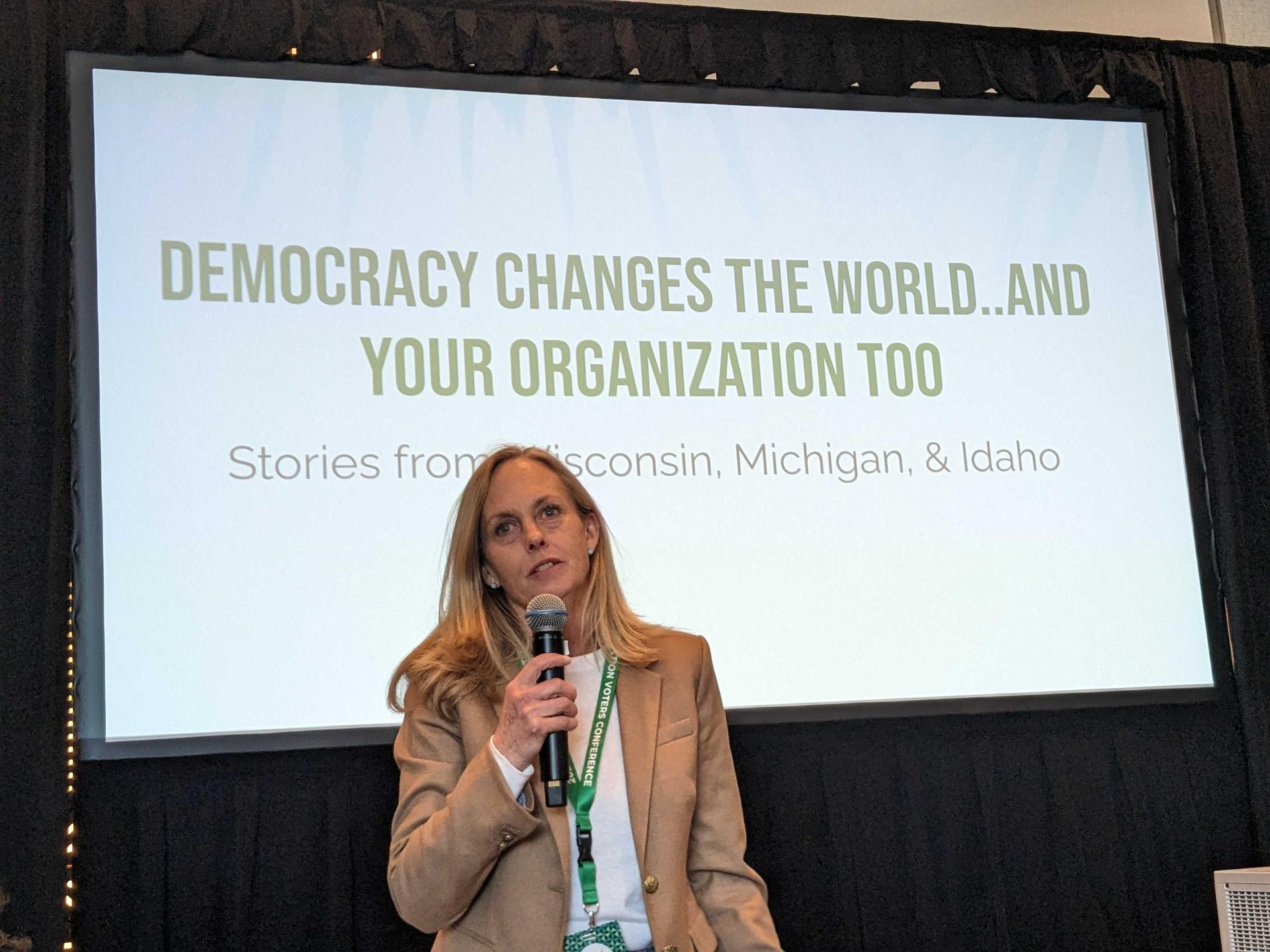 Michigan LCV Executive Director Lisa Wozniak speaking in front of a projector screen while holding a microphone. She has blonde hair and is wearing a tan jacket. 