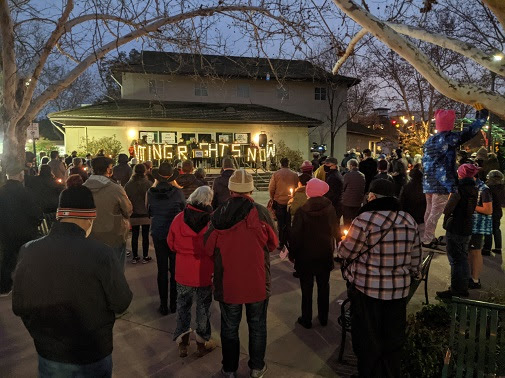 A crowd holds a candlelight vigil in Davis Central Park. A speaker stands on a stage in front of a sign made of LED letters reading "voting rights now" and addresses the crowd.