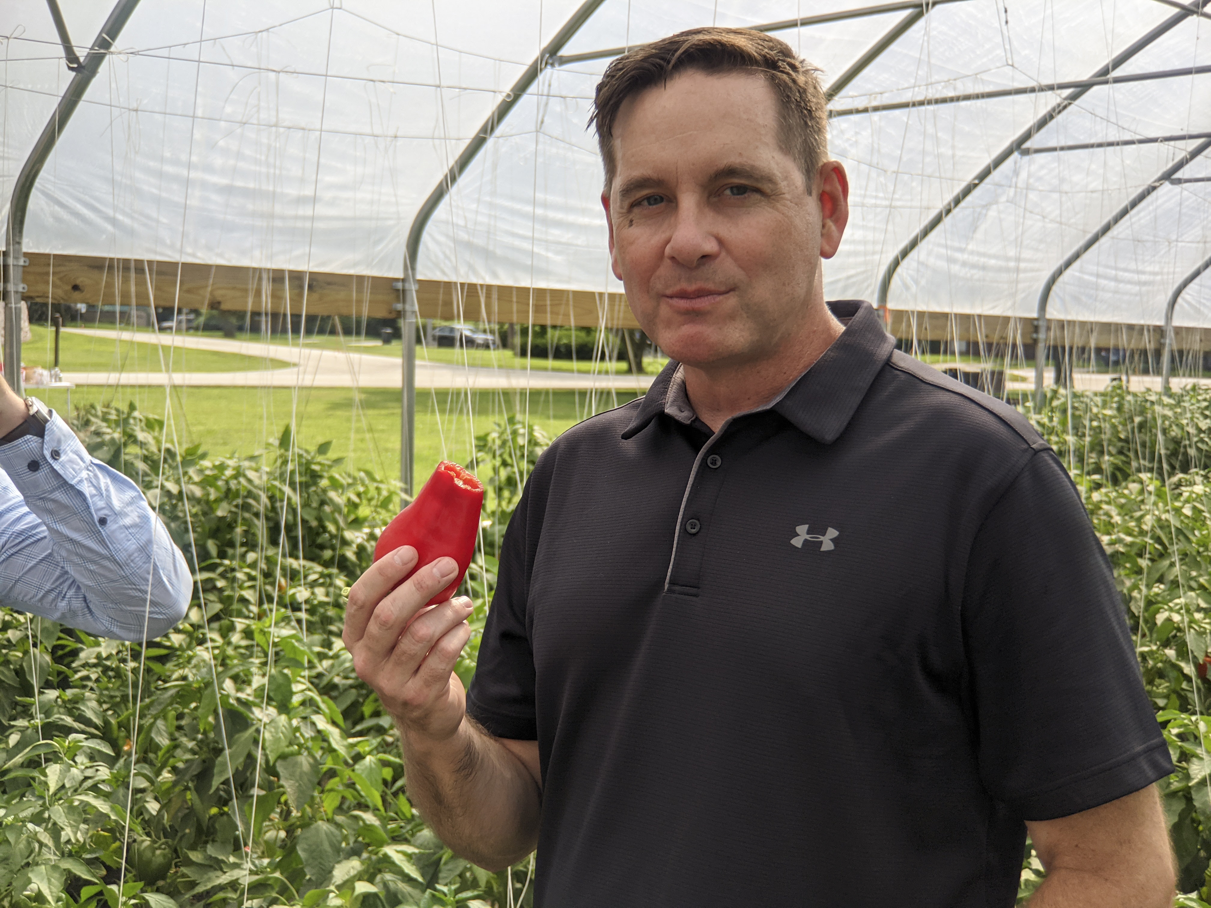 State Rep Tim Butler smiles at the camera after a tasty bite of sweet red pepper grown in the hoop house he's photographed in.