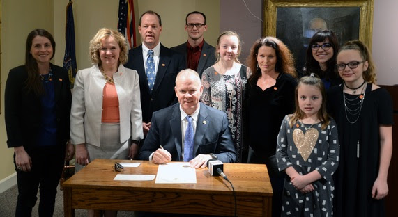 Wyoming Governor Matt Mead sits at a desk to sign the Random Acts of Kindness proclamation, surrounded by State Superintendent Jillian Balow, members of the Leadership Wyoming classmates that requested the proclamation, and three girls who raised money for Kindness Wyoming.