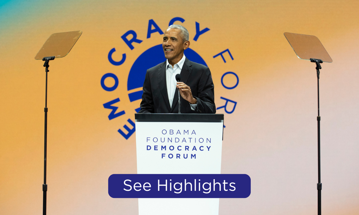 President Obama stands behind a podium as he speaks at the Obama Foundation Democracy Forum. The president has a medium skin tone and is wearing a black suit. In the background is a blue, green, and yellow gradient graphic that reads, “Democracy Forum.” At the bottom of the graphic is a blue rectangular button that reads, “see highlights.” 