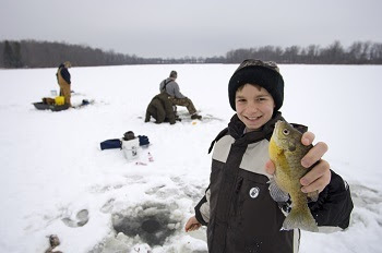 A young child with a beaming smile stands on a frozen lake and holds a panfish triumphantly.