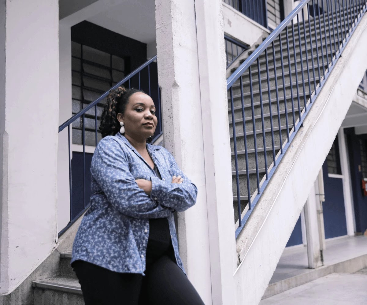 Priscila Cristina, ex-estudante do Prouni. Ela é uma mulher negra com cabelo castanho e cacheado. Priscila está encostada na viga de uma escada, com os braços cruzados. Ela veste camisa florida azul e calça preta