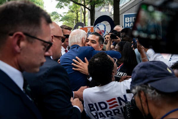 In the middle of a crowd of people, President Biden, seen from behind, embraces a man. A person wearing a Biden-Harris T-shirt is in the foreground.