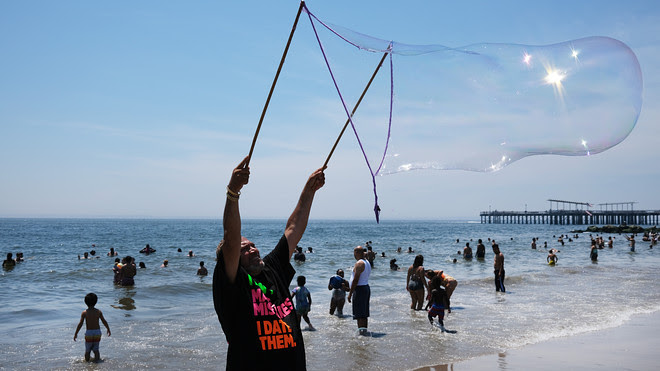 A picture of a man standing on the seashore blowing a massive bubble