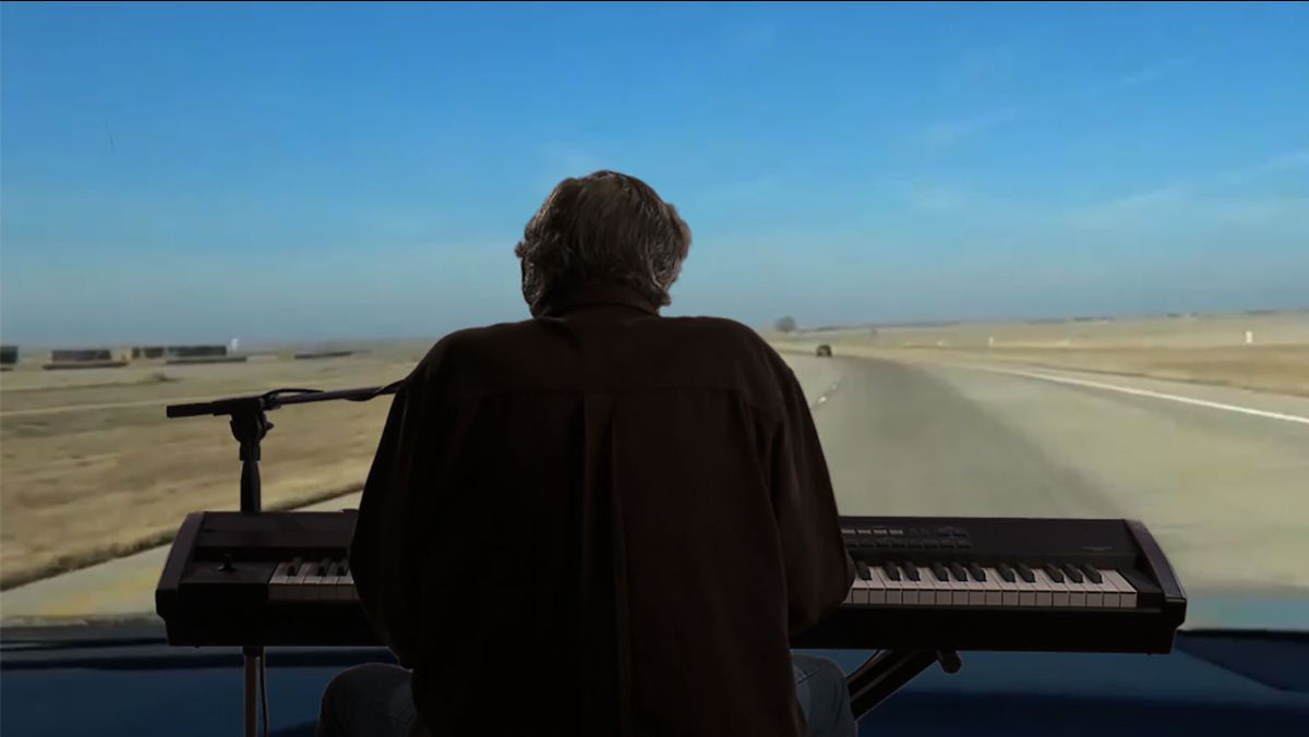 Man playing a keyboard and microphone seen from behind in front of video projection of desert road. 