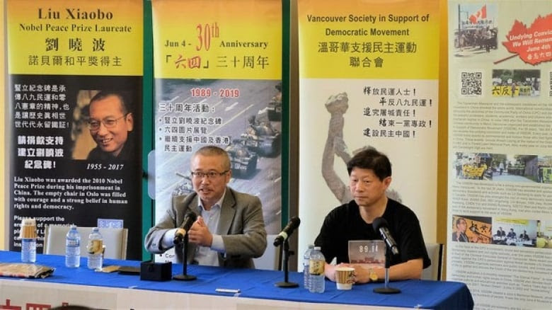 Li is sitting next to a man in front of yellow posters that say 'Vancouver Society in Support of Democratic Movement.'