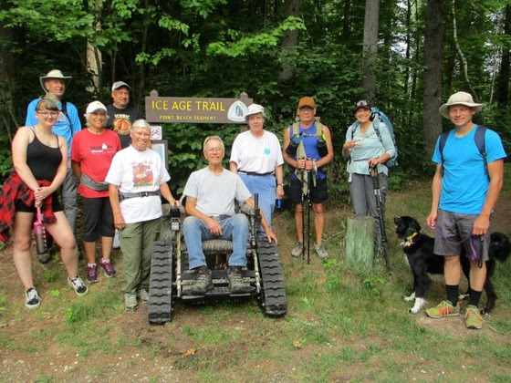 a group of people pose next to a man seated in an all-terrain wheelchair in front of an ice age trail sign