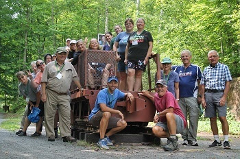 Men and women wearing shorts, T-shirts and other outdoor gear, stand on, around and in front of an old, orange mining flatbed