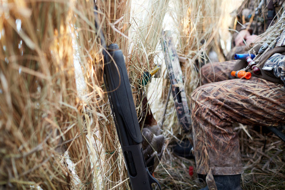 Hunters legs wearing camo are visible behind brush in a duck blind.