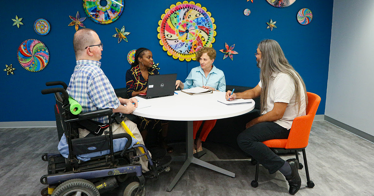 Four people are sitting at a roundtable taking notes on a notebook and computer.
