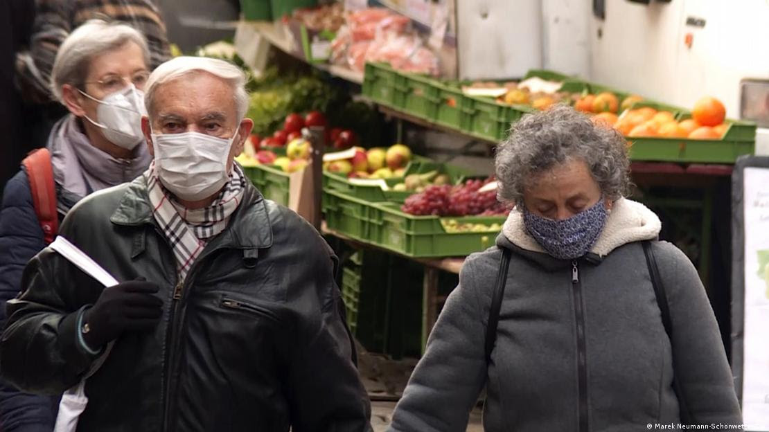 Três idosos, de máscara e roupas de frio, caminham em uma feira. Há frutas ao fundo. 