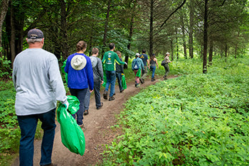 small group of adults in jeans and long-sleeved shirts hold bright green garbage bags as they walk single-file down a forested, dirt trail
