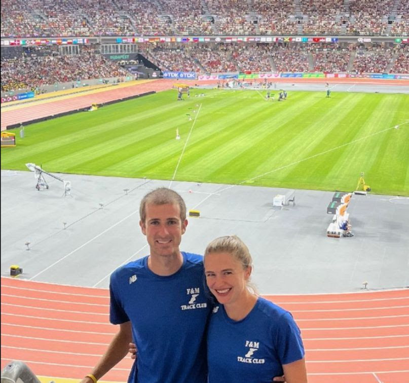 A white male with short brown hair and white female with brown hair in a ponytail stand in the foreground smiling. In the background is the World Championship Track in Budapest, Hungary.