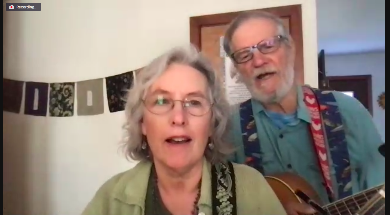 Two musicians with grey hair and glasses holding guitars and singing in their house