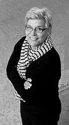 A black and white portrait of a light skinned woman with short white hair wearing thin glasses, a black shirt and pants, and striped scarf. The image is taken at a high angle, looking down at the woman who is looking up and smiling with her arms folded. 