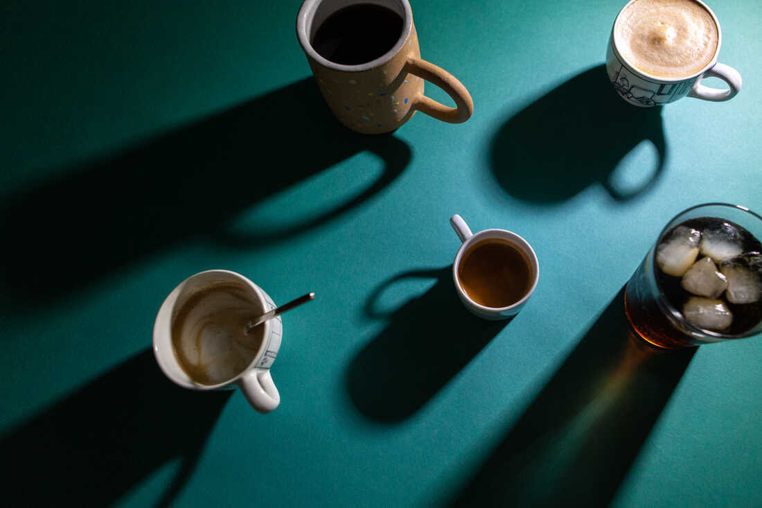 Image of five caffeinated drinks photographed from above on a dark green backdrop. A light from the right casts long shadows to the left of each beverage, including a latte, an espresso shot, a drip coffee, a dark brown soda and a cappuccino.